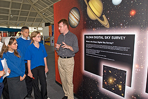 Dr. Bob Nichol talks to a group of students in front of 
    an SDSS museum exhibit, with pictures of planets on it